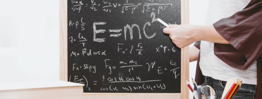 Female hand holding white cahlk and pointing to a small blackboard with calculations and E=MC2 equation on it